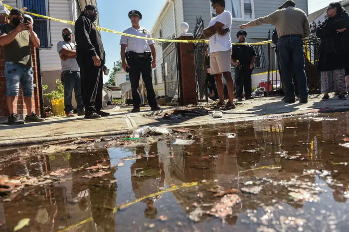 A puddle remains on the street, where a house has police tape with an officer standing and members of the media